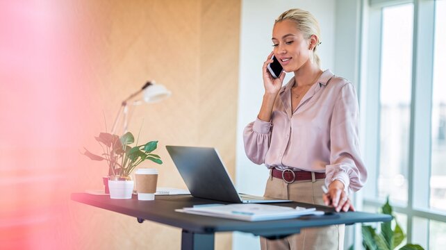 Confident Businesswoman on Phone Call in Modern Office. A confident businesswoman efficiently manages her workday, talking on her smartphone while working at a standing desk. 