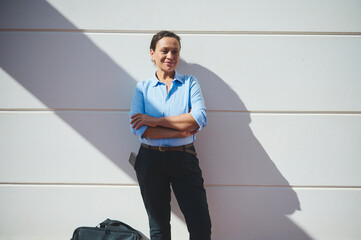 Professional woman in blue shirt standing confidently against a white wall with a shadow and...