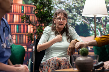 Caregiver chatting with elderly woman with disability during home visit, recommending medicine and supplements to help with recovery. Nurse and senior wheelchair user in a consultation.