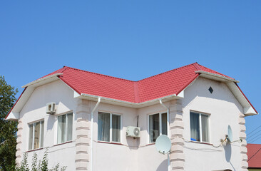 House with red lightweight metal roofing sheets, white plastic rain gutters, tv antenna, air duct and white painted walls against blue sky