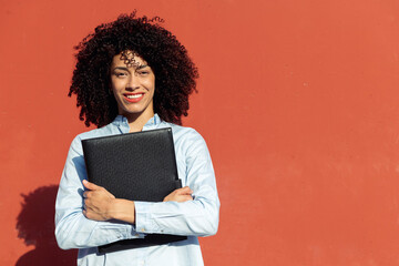 cheerful ethnic businesswoman with folder with documents standing on street near orange wall and looking at camera