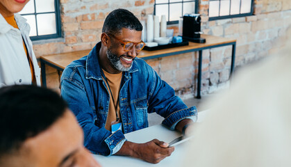Mature businessman in casual denim jacket smiling while using tablet during a relaxed meeting in a well-lit, collaborative environment