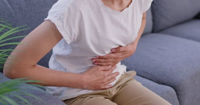 Woman sits on a couch at home, struggling with stomach pain and menstrual cramps. Discomfort of period aches, constipation, and the feeling of being sick and hurt during menstruation.