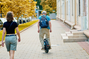 Senior man with backpack rides electric unicycle through urban streets. Man on electric scooter rides along sidewalk among other passers-by. Senior man riding an electric unicycle outdoors. © Tricky Shark