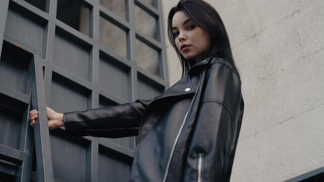 Stylish woman in a black leather jacket posing confidently near a modern architectural structure. Urban and edgy fashion shot capturing confidence and contemporary style. Confident Beautiful Woman