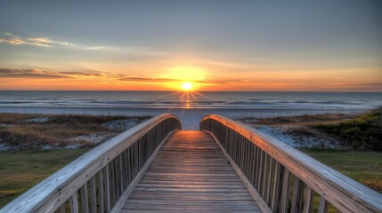 Obraz premium The wooden bridge leads toward the beach as the sun sets, filling the sky with warm hues of orange and pink while casting peaceful shadows on the sand.