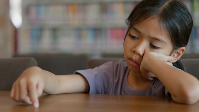 An Asian girl sits at a table in a library. She looks bored and lost in thought. Her disinterested expression shows her disinterest in the things around her.
