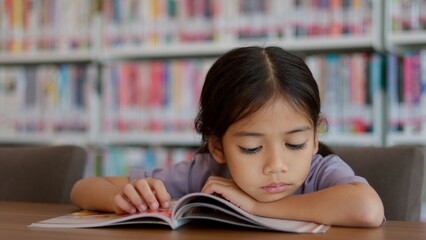 An Asian girl is reading a book in a library, looking serious as she focuses on the contents.