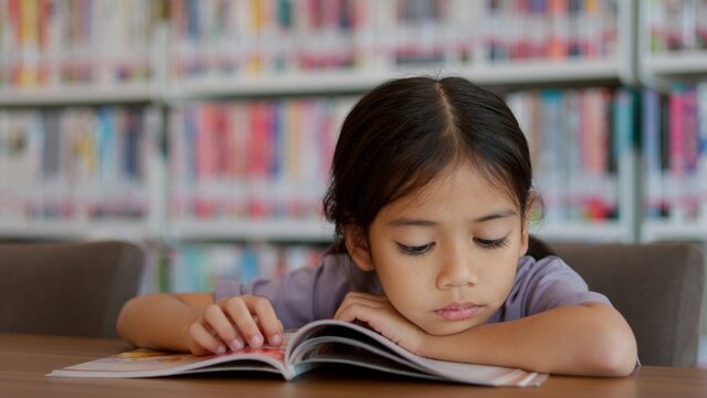 An Asian girl is reading a book in a library, looking serious as she focuses on the contents.