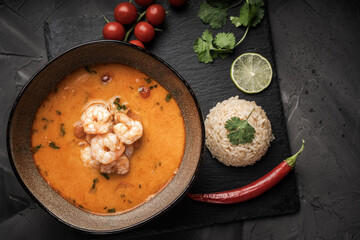 Tom Yam Kung Spicy Thai soup with shrimp in a bowl on a dark background and red pepper, cilantro rice on a black mica board. top view
