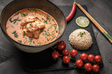 Tom Yum Kung Spicy Thai soup with shrimp in a bowl on a dark background and red pepper, rice and lime with cilantro and chopsticks on a black mica board. Copyspace
