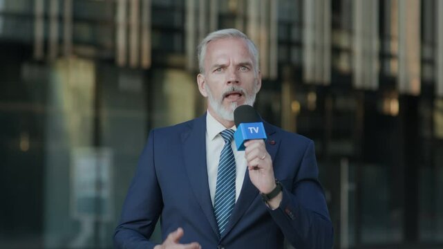 Medium shot of adult Caucasian male news presenter wearing blue suit walking with microphone with blue TV logo towards camera while covering report outside modern glass building
