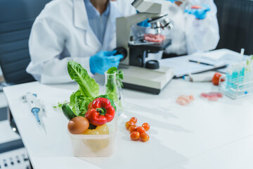 Two Asian women in a lab work on food research, using a microscope, petri dish, and test tubes filled with chemical solutions. They study vegetables, pork, and plants for GMO traits and nutrition.