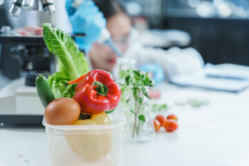 Two Asian women in a lab work on food research, using a microscope, petri dish, and test tubes filled with chemical solutions. They study vegetables, pork, and plants for GMO traits and nutrition.