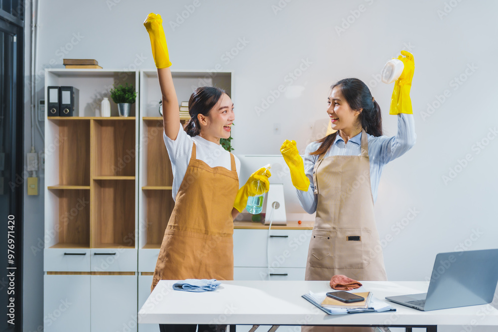 Wall mural two asian housekeepers, wearing aprons and rubber gloves, happily clean an office. they sweep the fl