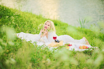 A beautiful blonde woman with a bouquet of flowers is lying on a blanket in a meadow near a river on a hill. Picnic