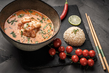 Tom Yum Kung Spicy Thai soup with shrimp in a bowl on a dark background and red pepper, rice and lime with cilantro and chopsticks on a black mica board. Copyspace