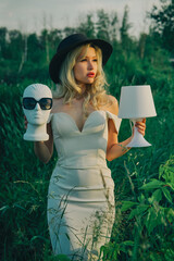Beautiful fashionable girl with a hat and a lamp in her hands, holding the head of a mannequin wearing glasses in nature near a pond