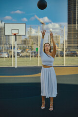 Glamorous woman in white dress playing basketball on court. Sportswoman throwing a ball into a basket. Concept sport, fashion.