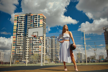 Confident young female holding a ball in her hands on a basketball court. Lady in white dress with slit and heeled shoes. Fashion, sports concept