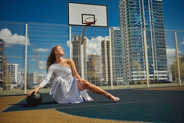 Athletic, woman in white dress poses on basketball court. Glamorous lady holding a black ball. Fashion, sport concept