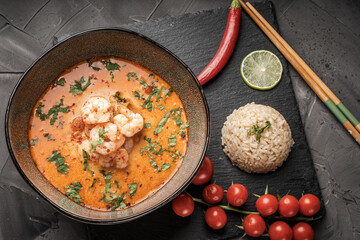 Tom Yum Kung Spicy Thai soup with shrimp in a bowl on a dark background and red pepper, rice and lime with cilantro and chopsticks on a black mica board. Copyspace