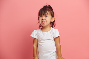 Little Asian baby girl shows tongue wears t shirt isolated over pink background. Three years old offended and capricious child.