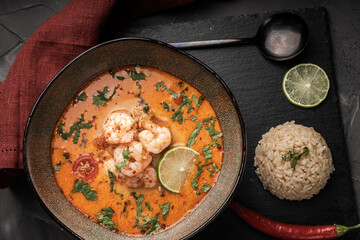 Tom Yam Kung Spicy Thai soup with shrimp in a bowl on a dark background and red pepper, rice and lime with cilantro and a spoon on a black mica board.