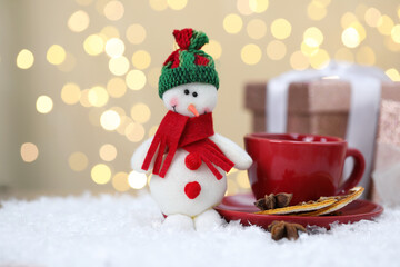 Funny snowman figure, cup, dried orange slices and cinnamon on artificial snow against blurred lights
