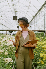 Woman in casual attire analyzing flower growth in greenhouse environment, holding notepad and pen while observing plants around her, yellow flowers dominating the scene