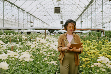 Woman standing among blooming flowers in large greenhouse, wearing hat and glasses, writing on...