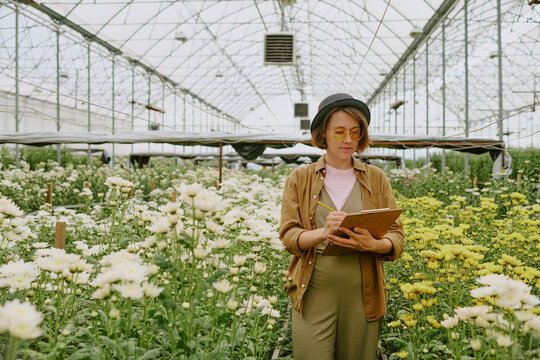 Woman standing among blooming flowers in large greenhouse, wearing hat and glasses, writing on clipboard with a focused expression, plants thriving around