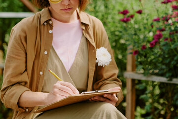 Person sitting in garden, writing notes on clipboard with yellow pencil Flowers blooming and...