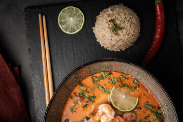 Tom Yum Kung Spicy Thai soup with shrimp in a bowl on a dark background and red pepper, rice and lime with cilantro and chopsticks on a black mica board. Copyspace