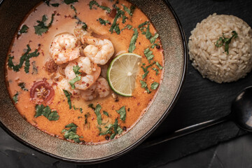 Tom Yam Kung Spicy Thai soup with shrimp in a bowl on a dark background and red pepper, rice and lime with cilantro and a spoon on a black mica board.