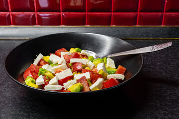 Fresh red tomato, avocado and bryndza or feta salad with olive oil in black dish with a spoon close-up side view, healthy eating.