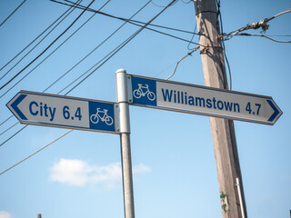 The road sign in Melbourne points to two destinations: City, 6.4 kilometers away, and Williamstown, 4.7 kilometers away. Both routes are suitable for cyclists, with the bicycle symbols on the sign