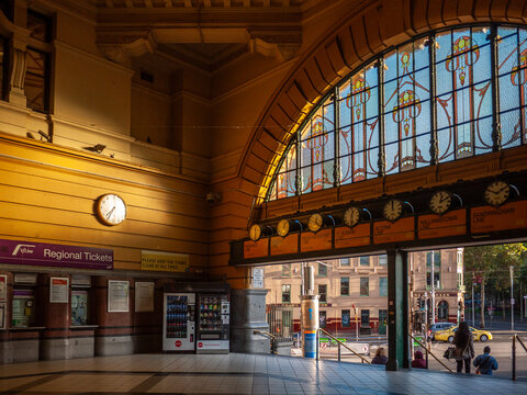 The interior of Flinders Street Station in Melbourne, Australia, with its grand arched entrance, is illuminated by sunlight and adorned with ornate details of a row of vintage clocks.