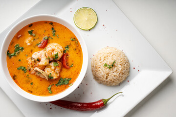 Tom Yam Kung Spicy Thai soup with shrimp and cilantro in a white bowl on a light background with rice and red hot pepper, lime and chopsticks on a square plate. Top view, close-up. Copyspace.