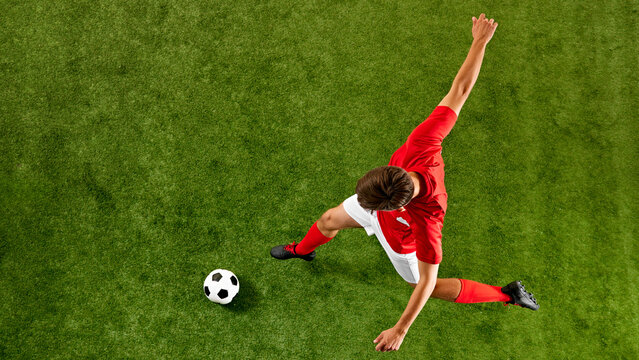 Athletic footballer in red-white, performing swift volley kick during football training on green pitch. View from high angle. Concept of championship, sport events, final game. Ad