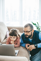 A father and his little daughter lying on a couch, focusing together on a laptop screen, possibly for studying or leisure