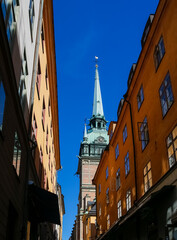 Obraz premium Tall spire of a historic clock tower of Tyska kyrkan or German Church stands proudly between two narrow streets lined with vibrant buildings, bathed in sunlight against a brilliant blue sky.