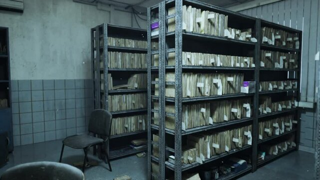 The abandoned hospital features decaying shelves filled with old medical records, revealing the neglect and deterioration of a once functional healthcare facility. A chair sits forlornly nearby.
