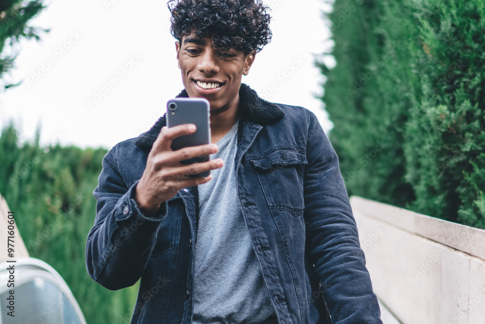 Poster happy hipster guy going down at city escalator and using modern cellphone gadget for online communic