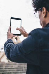 Back view of millennial male tourist using modern blank touch pad with copy space for advertising for clicking photos of city landscape in historic center, travel man photographing buildings