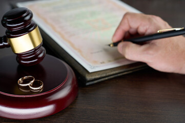 Man hand signing divorce contract, close-up. Wedding rings with marriage contract and judge gavel on a table