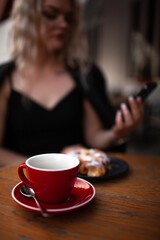 A stylish girl of twenty-five years old sits in a cafe on the outdoor terrace, drink coffee and eat croissant, the woman is wearing a black dress, jacket and glasses,