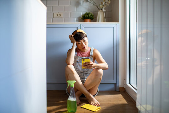 Young woman cleaning kitchen while checking her smartphone at home