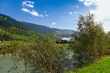 a steam train driving at a sunny day at a river in the austrian mountains in pinzgau