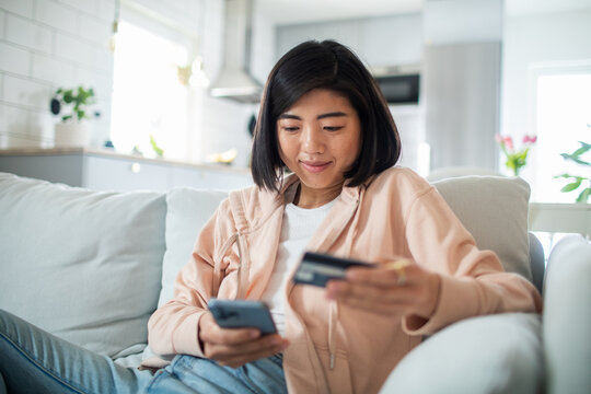 Young Asian woman holding credit card and smartphone on home sofa